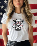 White t-shirt with a cartoon Highland cow wearing sunglasses and holding an American flag.